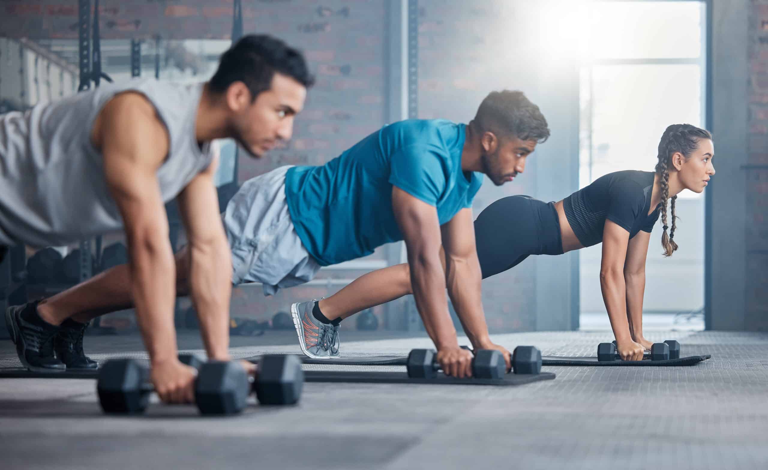 Trois athlètes, deux hommes et une femme, maintiennent une position de planche haute avec haltères dans un gymnase moderne et lumineux.