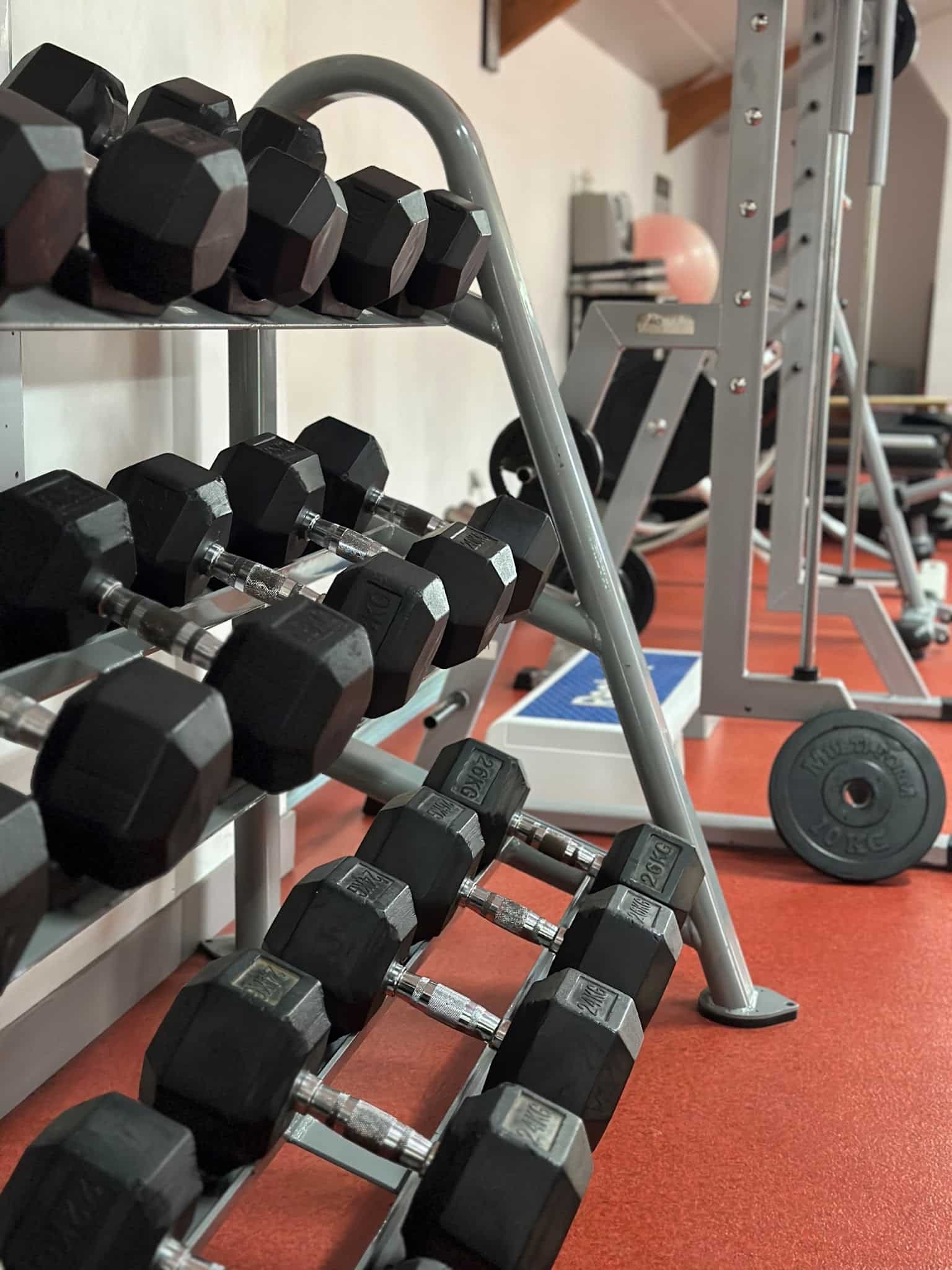 Haltères Musculation : Rack et Équipement de Fitness Rangement de haltères hexagonaux noirs, pesant jusqu'à 26kg, dans une salle de sport au sol rouge, avec un rack de musculation en arrière-plan.
