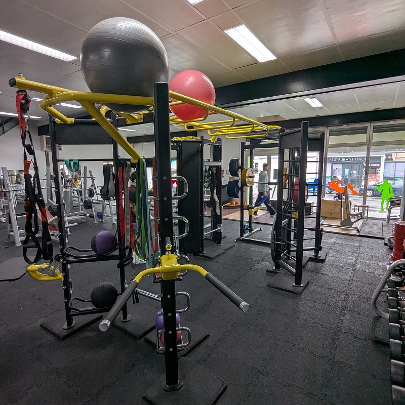 Salle de sport : musculation, accessoires et vitrine Intérieur d'une salle de sport avec équipement de musculation jaune et noir, ballons de stabilité, sangles TRX et un homme marchant devant une vitrine affichant "RESTAURANT ITALIEN".