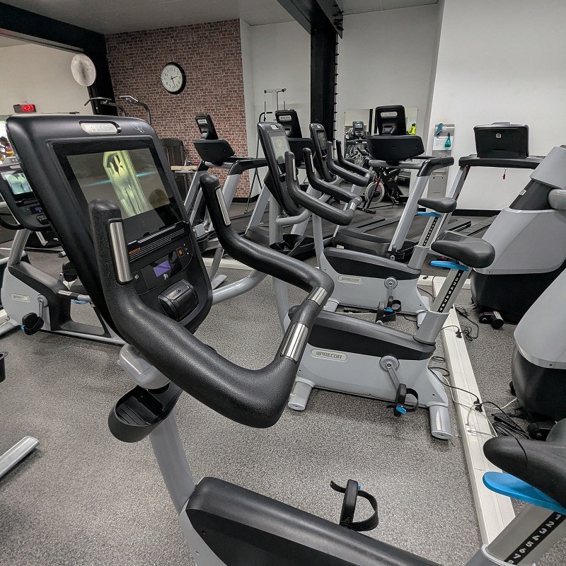 Vélos Stationnaires Precor dans une Salle de Sport Moderne Intérieur de salle de sport avec vélos stationnaires Precor, l'écran du vélo au premier plan montre un affichage jaune. Une horloge murale indique environ 10h10.