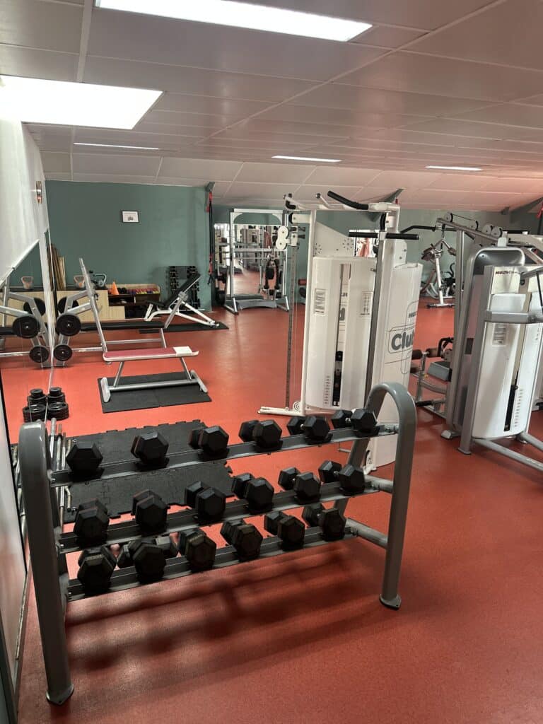 Équipement de musculation et fitness en salle de sport Salle de sport équipée : rack d'haltères, bancs et machines MultiForm sur un revêtement de sol rouge.